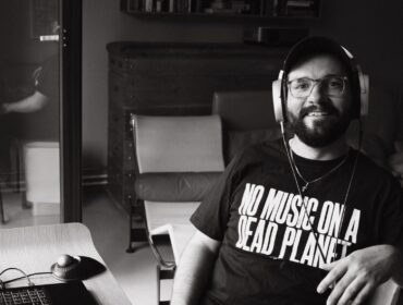 Flo Siller wearing headphones at his workstation, his shirt reads No Music on a Dead Planet